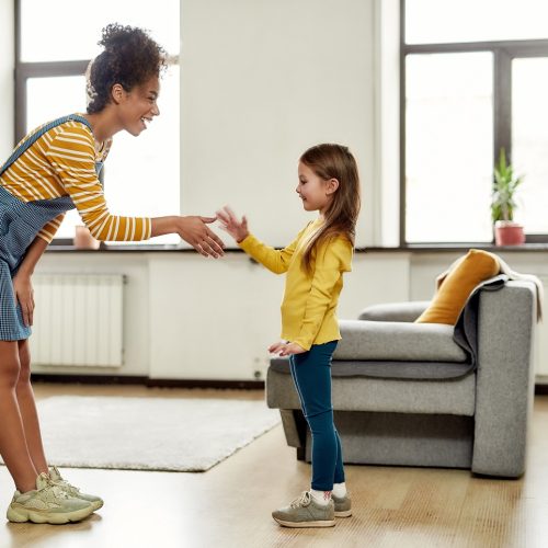 Full length shot of african american woman baby sitter get acquainted with caucasian cute little girl. They are standing indoors and talking. Babysitting, nanny concept. Horizontal shot. Side view